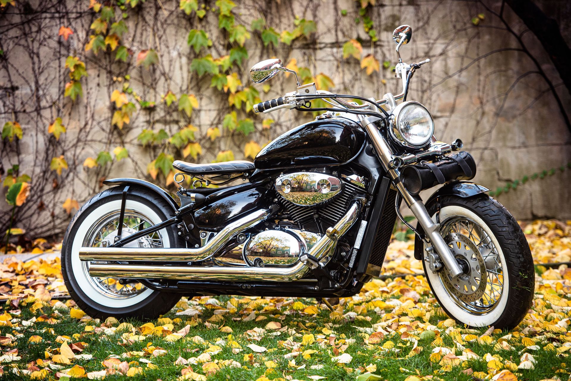 Classic vintage motorcycle parked on fallen autumn leaves with ivy-covered wall backdrop.